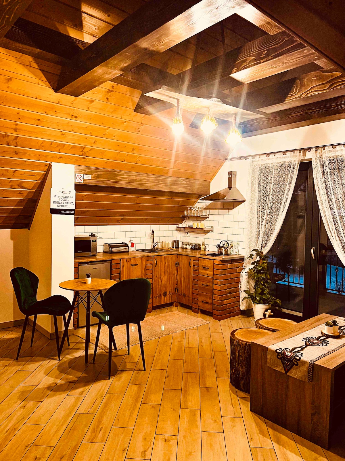 A rustic kitchen area showcases wooden cabinetry and modern appliances, including a microwave and a stove. A round table with two chairs is positioned nearby. Natural light filters through a large window, complemented by sheer curtains, enhancing the warm wood accents of the vaulted ceiling.