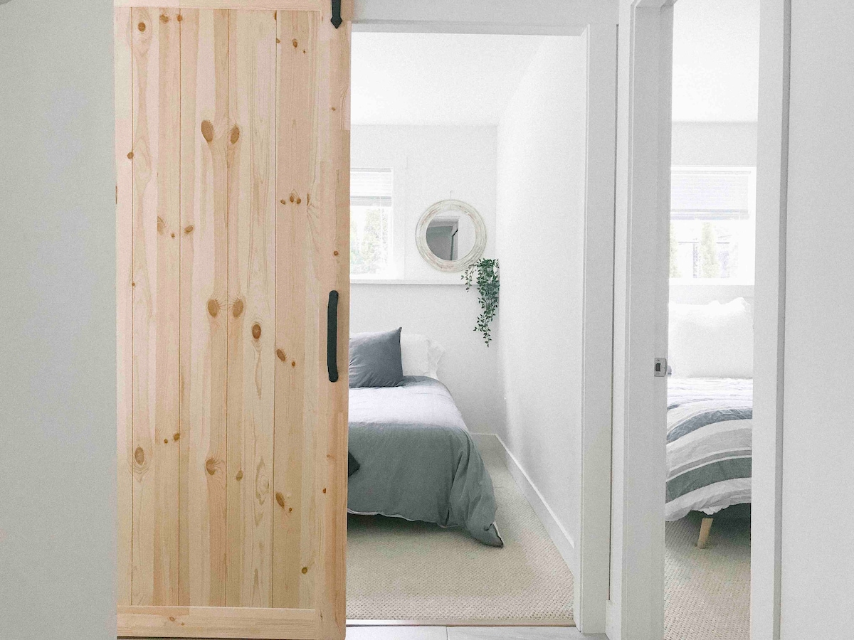 A light and airy entrance showcases a wooden sliding door leading into a room with a spacious bed, adorned with soft grey bedding. Natural light floods in through a nearby window, illuminating the minimalist décor, which includes a round mirror and a touch of greenery.
