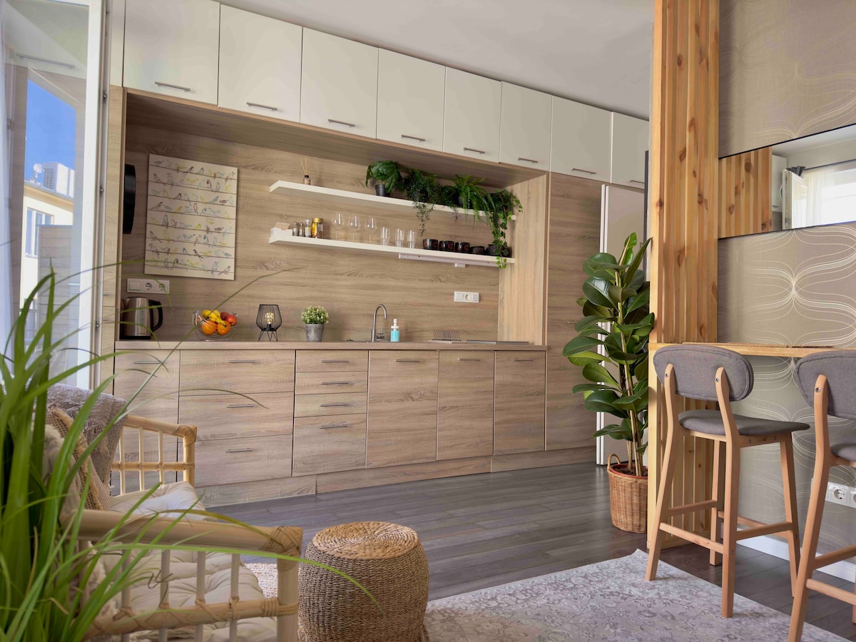 The modern kitchen features wooden cabinetry and open shelving, showcasing glassware and decorative plants. A counter with bar stools provides a casual dining space. Natural light enters through large windows, complemented by indoor greenery, creating a functional yet inviting environment.