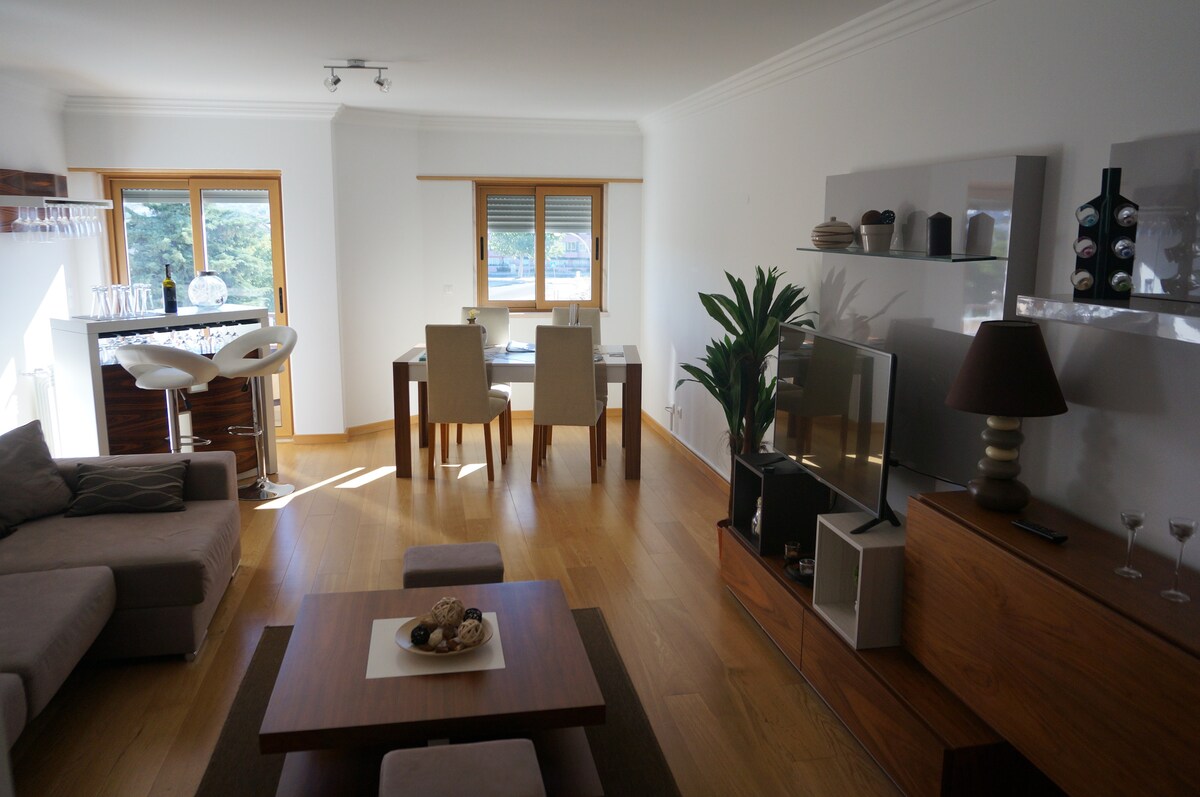 A spacious living area is presented, featuring a comfortable sofa and a modern coffee table. Dual windows allow natural light to brighten the space, complemented by stylish dining and bar seating. Decorative elements, including plants and a sleek television unit, enhance the inviting ambiance.