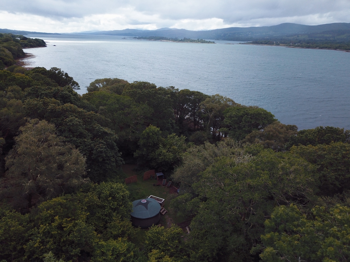 An aerial view captures the lush green surroundings of the glamping site, with the spacious Mongolian yurt centrally located. The tranquil waters of Kenmare Bay stretch out in the background, framed by dense woodlands and distant hills under a partly cloudy sky.