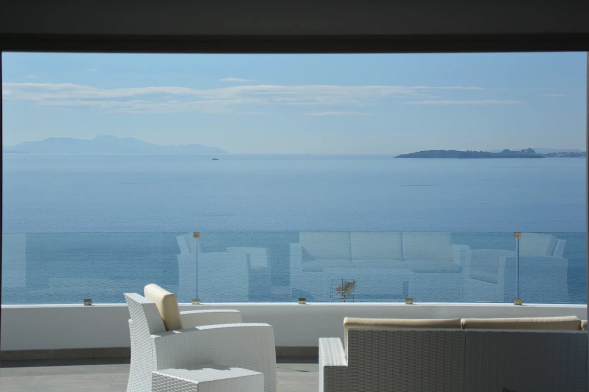A spacious outdoor seating area is visible, featuring white wicker chairs and a sofa. The glass railing offers unobstructed views of the calm sea and distant landmasses, creating an open and serene atmosphere. Light clouds are present in the clear blue sky.