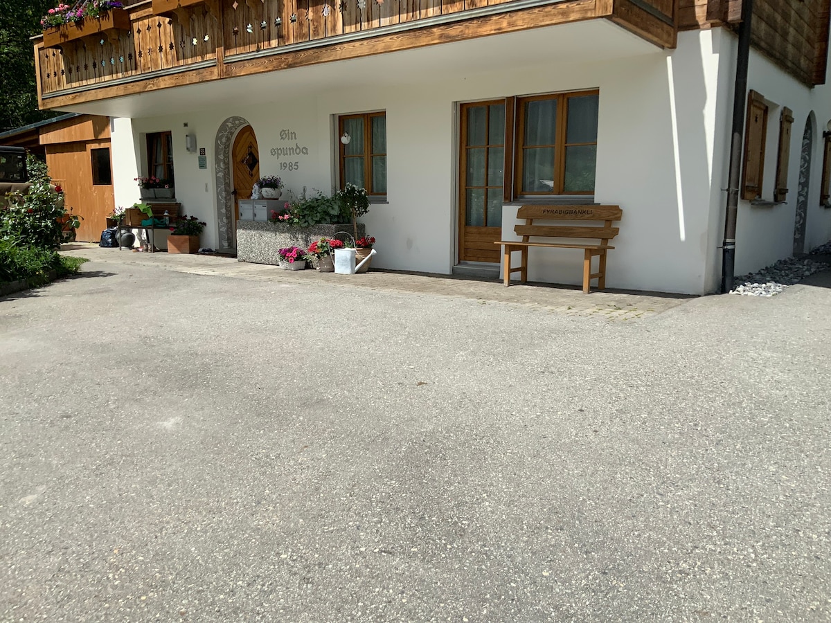 A charming exterior view of a house displays a well-maintained facade with wooden accents. Flower pots are arranged along the entrance, and a wooden bench is positioned in front. The paved area is clear, providing ample space in front of the building.