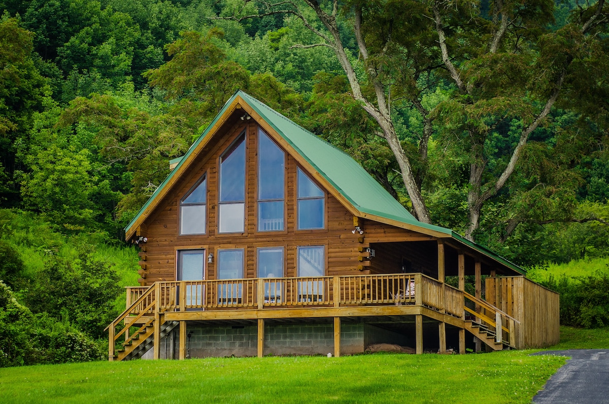 A log cabin features a triangular roof and large glass doors that provide a view of the surrounding greenery. The wraparound deck is accessible by stairs, offering an inviting outdoor area. Lush trees frame the cabin, enhancing the tranquil setting.