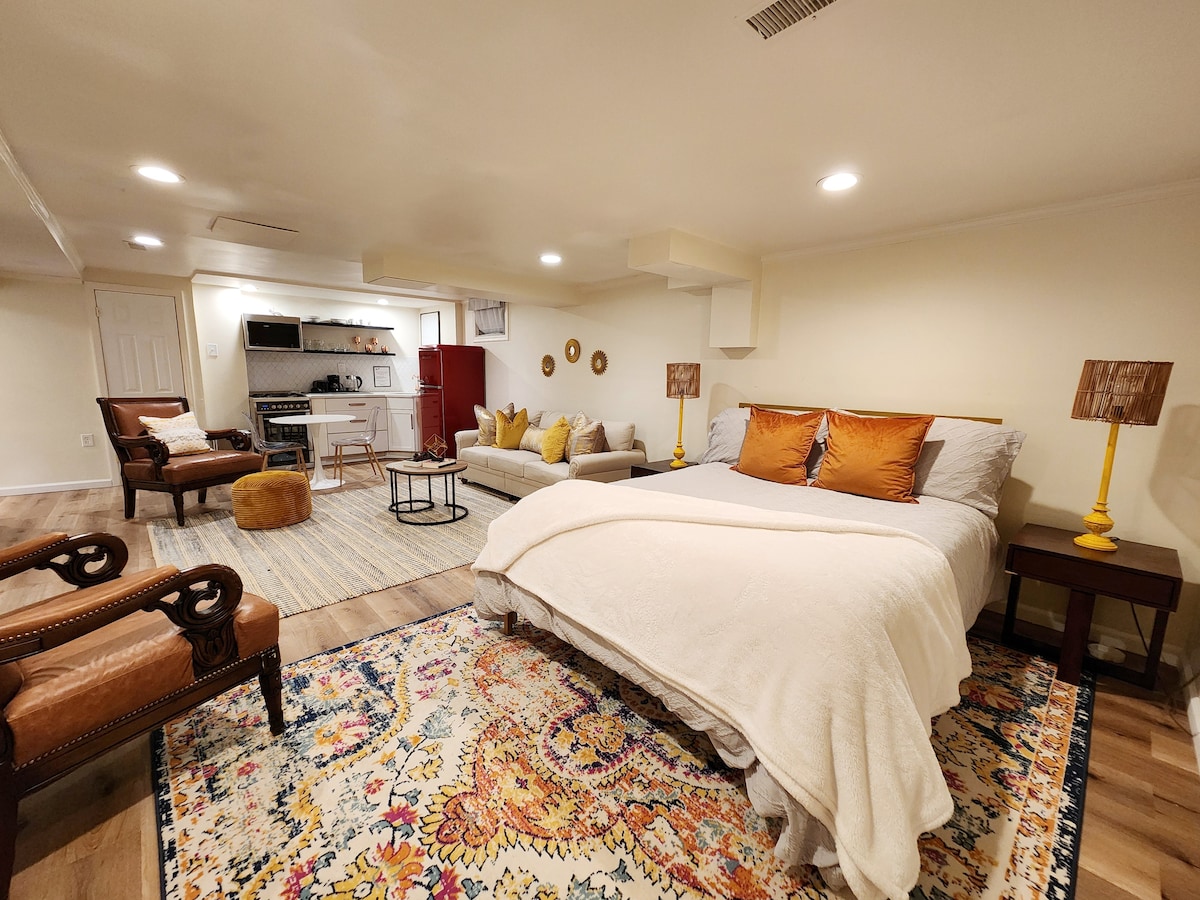A spacious studio with a large bed dressed in neutral linens, accentuated by decorative pillows. A cozy seating area with a sofa and chairs is complemented by a vibrant area rug. Soft lighting creates a warm ambiance, and a kitchenette is visible in the background.