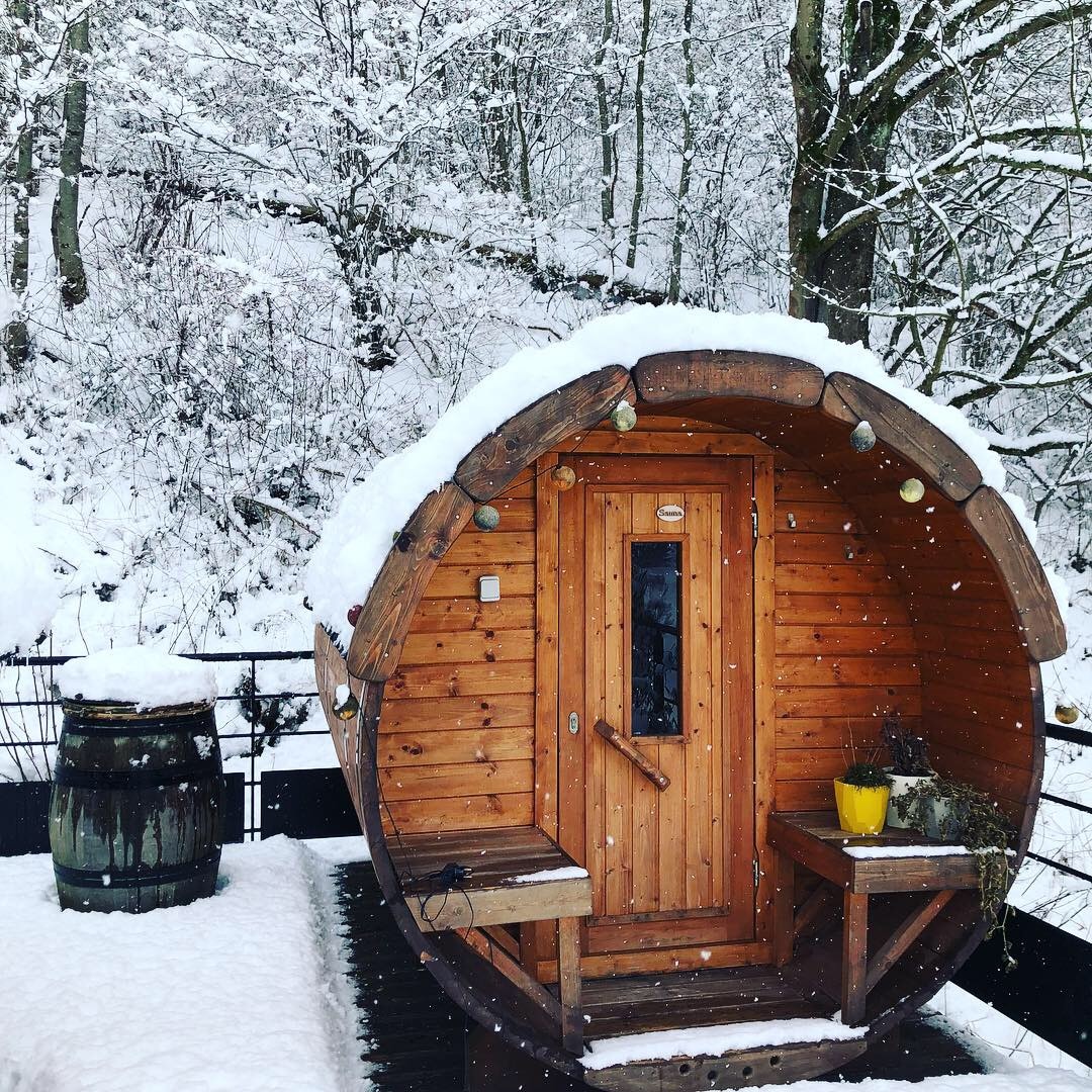 A wooden sauna is nestled in a snowy landscape, featuring a rounded design with a door and small window. Snow covers the roof and surrounding area, while a barrel sits nearby, adding to the serene winter setting.