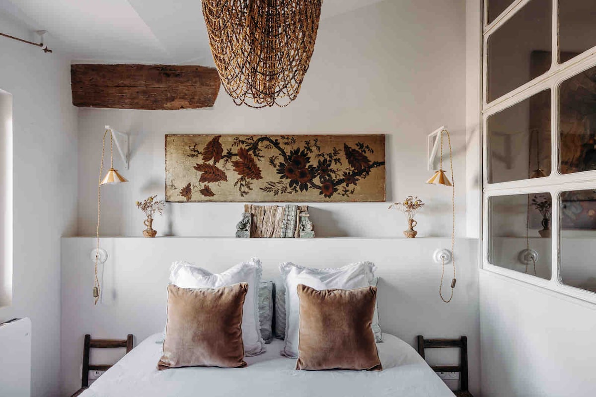 A serene bedroom features a spacious bed adorned with plush pillows. Unique lighting fixtures are mounted on the walls, and an intricately designed textile hangs above the headboard. Natural light filters through the window, highlighting the blend of modern and vintage decor.