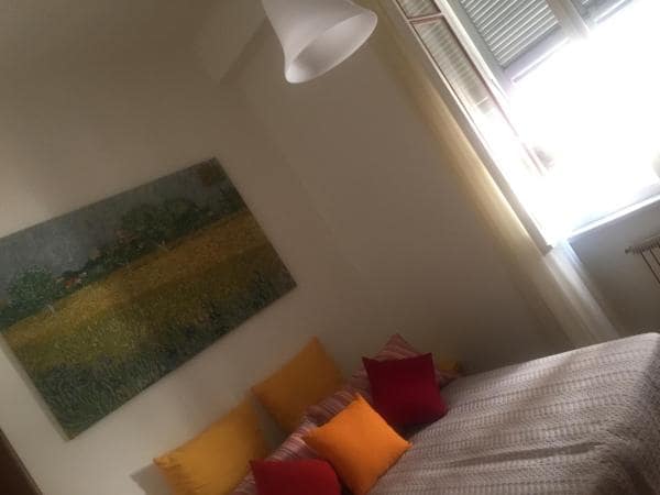 A bright bedroom features a spacious bed adorned with colorful cushions in shades of orange and red. Natural light filters through the window, illuminating a large wall painting depicting a serene landscape. The overall design conveys a comfortable and inviting space.