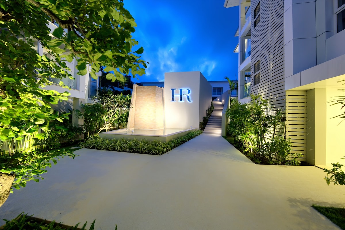 The entrance of Horizon Residence is presented, highlighting a water feature and illuminated 'HR' signage. Lush greenery surrounds the pathway, with soft lighting accentuating the foliage. The well-maintained walkway leads to the buildings, creating an inviting approach to the condominium.