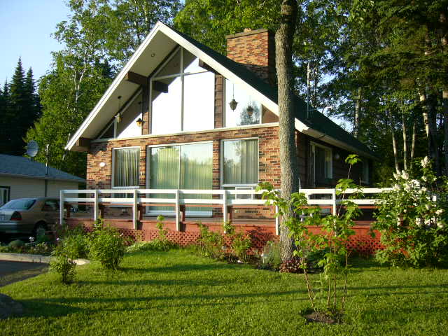A Swiss-style chalet is set amidst lush greenery, featuring large glass windows that allow natural light to permeate the interior. A wooden deck extends in front, complemented by a manicured lawn and surrounding trees, presenting a welcoming exterior.