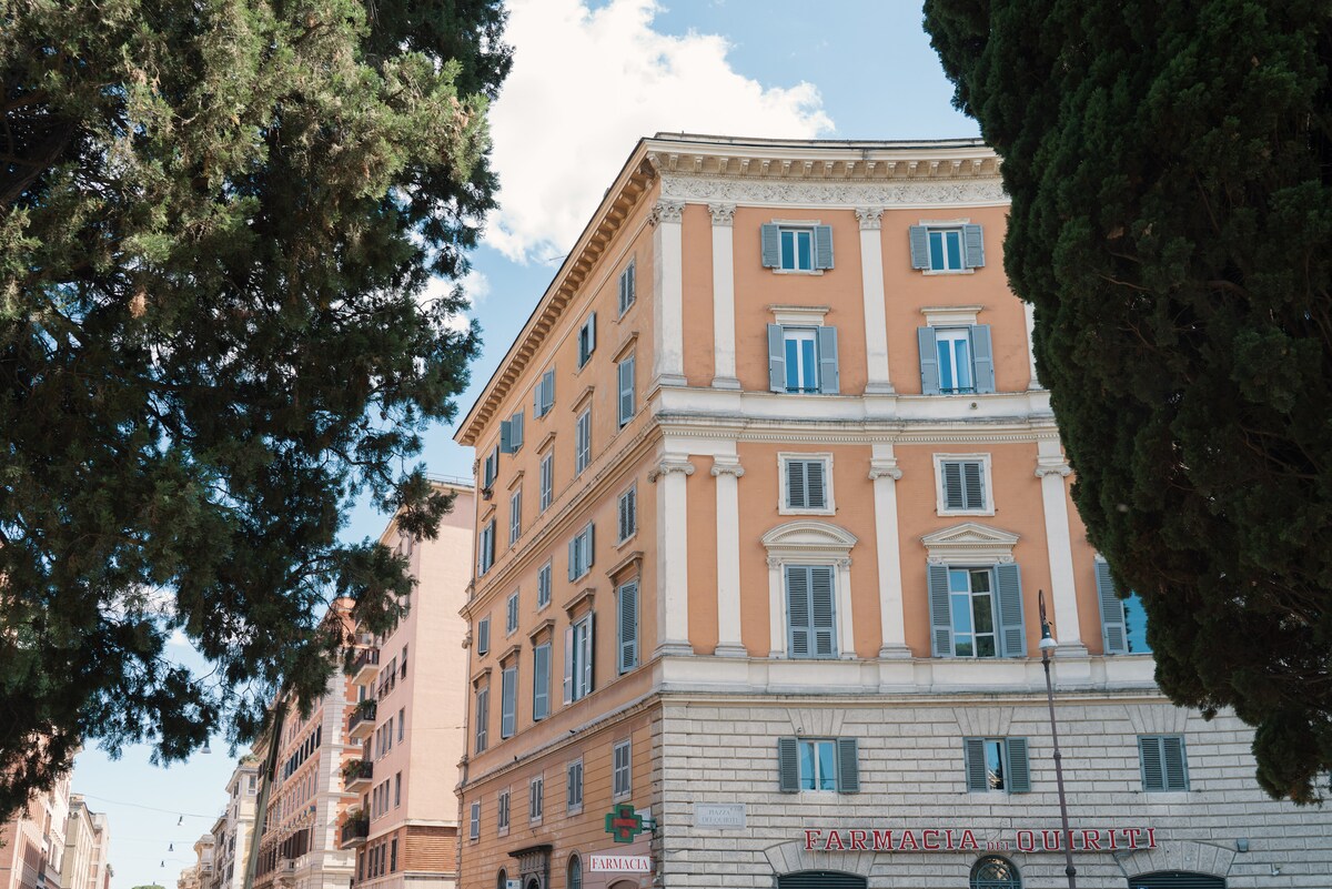 The exterior of a historic building is depicted, showcasing elegant architectural details, including large columns and vibrant orange walls. Lush green trees frame the image, adding a natural element to the urban landscape.