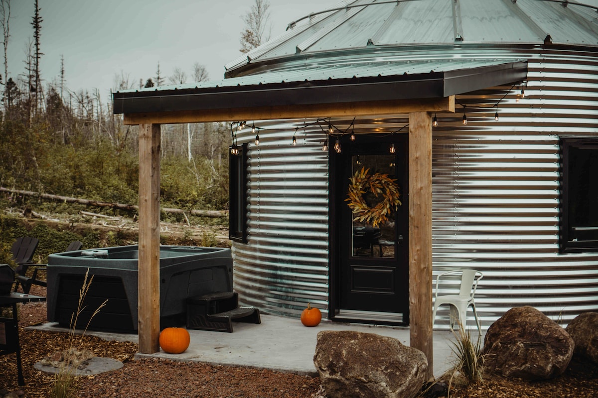 The exterior features a round, metal-clad structure with large windows and a black door. A private spa is positioned next to the entrance, flanked by decorative pumpkins. String lights are suspended above a sheltered entry area, providing a cozy touch to the natural surroundings.