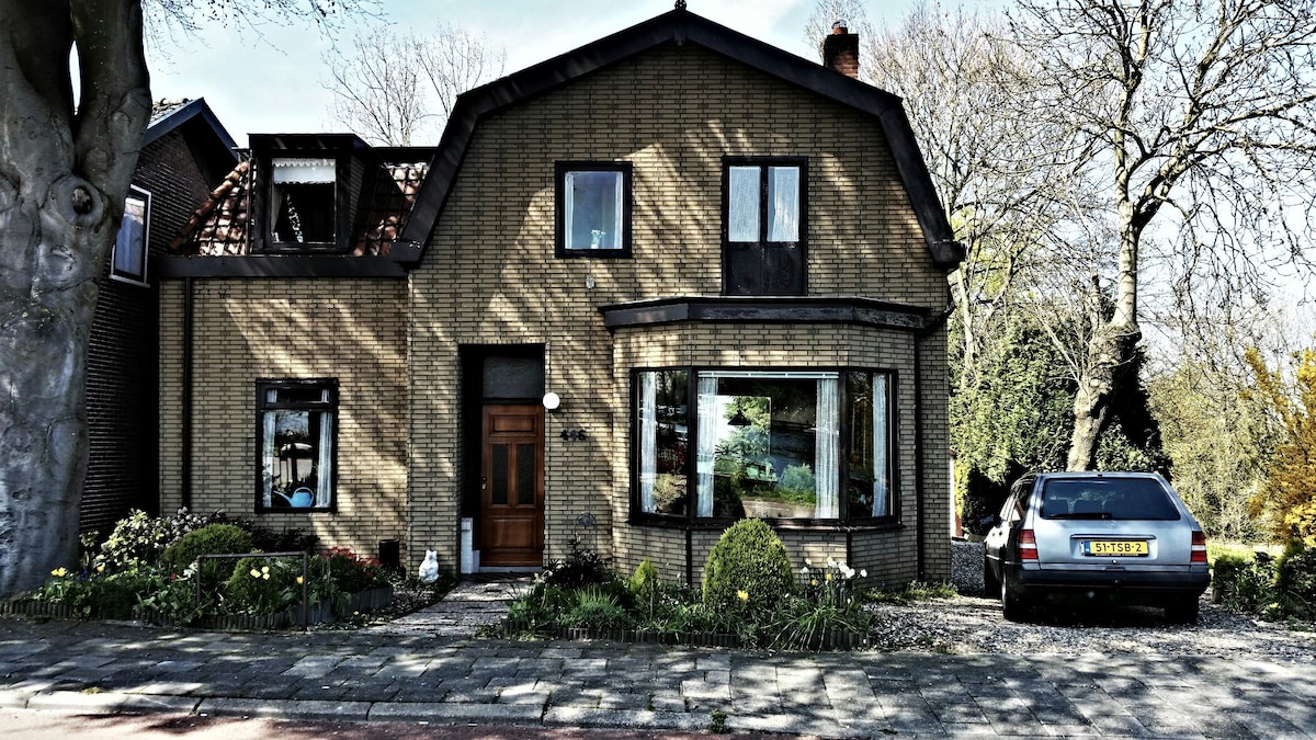The exterior of a Dutch house features a unique architectural design with a steeply pitched roof. Large windows offer views of the surrounding greenery, and a well-maintained garden surrounds the front entrance, creating a welcoming atmosphere. A parked car is visible in the driveway.