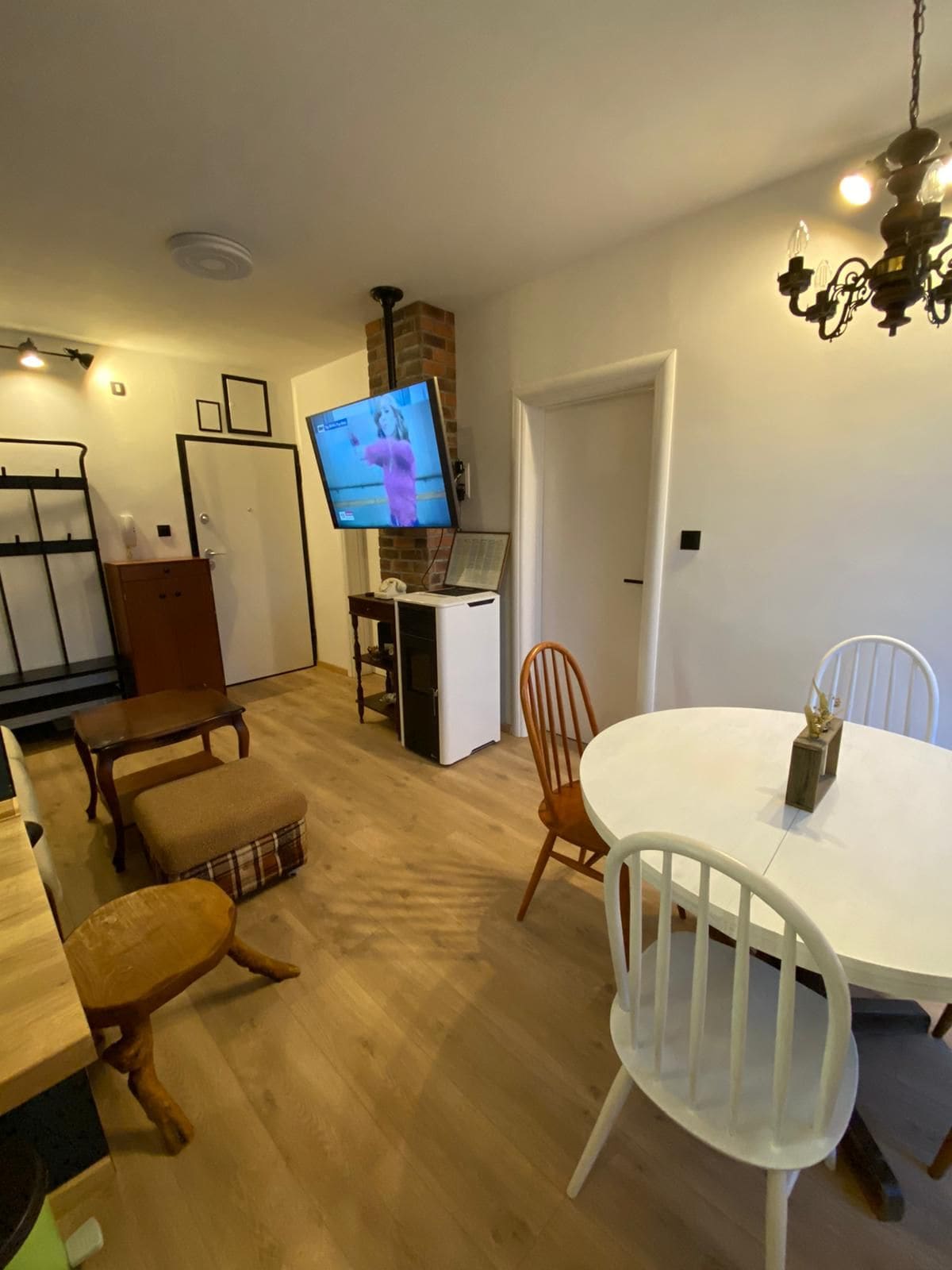The dining area features a round table surrounded by two wooden chairs and a bench with a woven seat. A television is mounted on the wall, while light from a chandelier adds illumination to the space. A small kitchen area and an entryway are visible.
