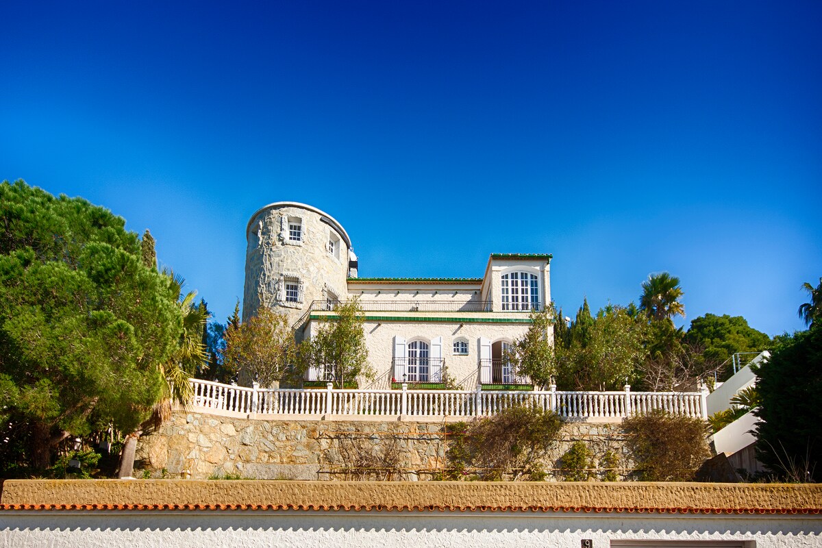 A charming villa is situated on a hillside, showcasing a blend of stone and white architecture. Lush greenery surrounds the property, with a spacious terrace visible in front. The clear blue sky serves as a backdrop to this inviting home.