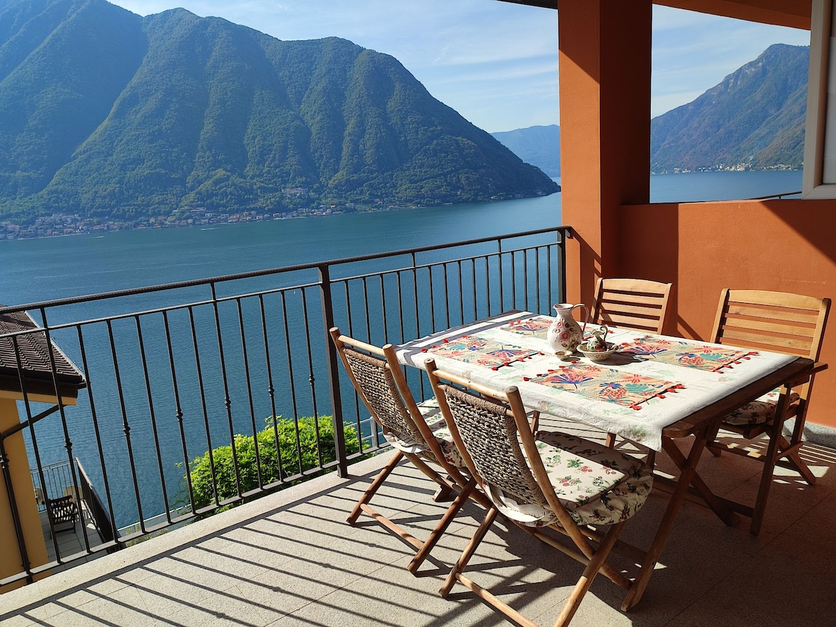 An outdoor terrace features a spacious table surrounded by wooden chairs, with a colorful tablecloth. The panoramic view showcases a tranquil lake bordered by mountains, enhancing the inviting setting for meals or relaxation.