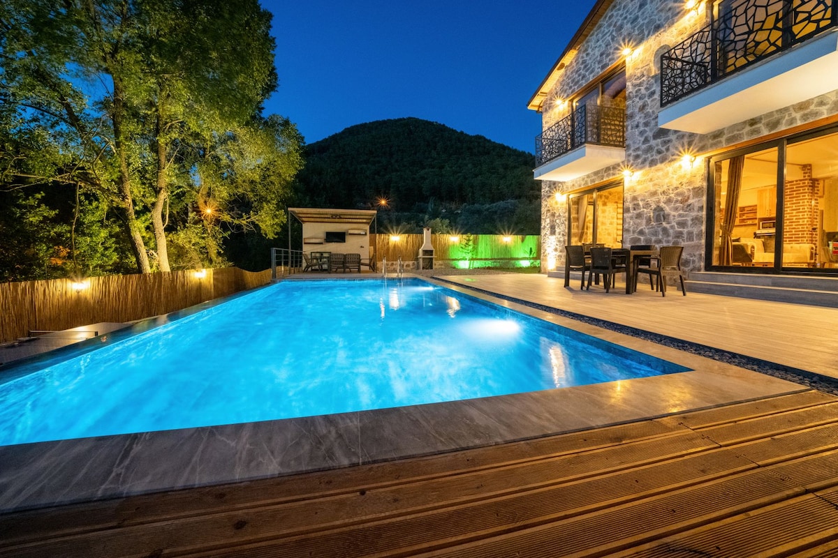 A well-lit outdoor area features a spacious swimming pool surrounded by wooden decking. The pool's turquoise water is softly illuminated, reflecting the evening sky. The stone villa stands nearby, with outdoor seating visible on the patio, framed by greenery and gentle hillside slopes.