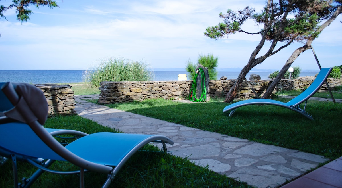 A serene outdoor space is showcased, featuring two lounge chairs positioned on green grass. A stone wall borders the area, overlooking the tranquil sea. Lush vegetation and a clear sky enhance the peaceful coastal ambiance.