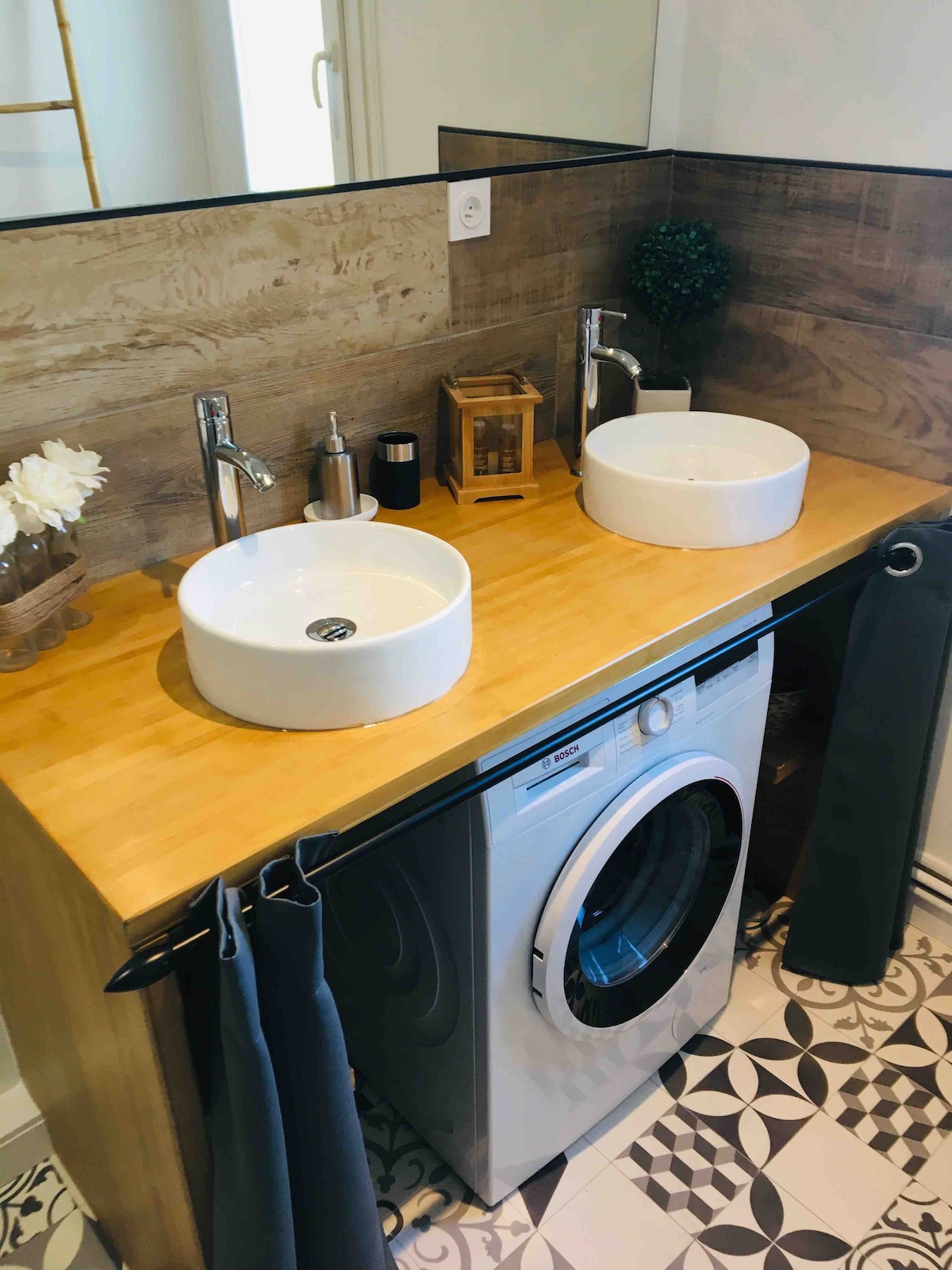 A modern bathroom features dual round sinks set on a wooden countertop. A washing machine is integrated below, with a decorative plant and a lantern adding detail. The floor is adorned with patterned tiles, enhancing the overall aesthetic.