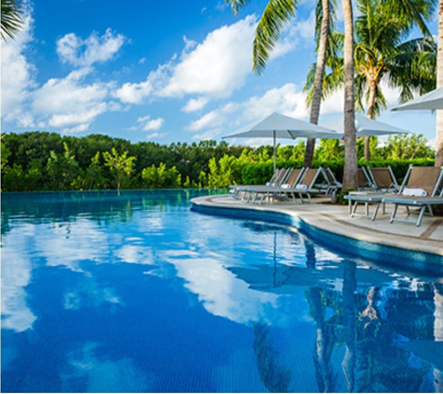 A serene swimming pool is bordered by palm trees and sun loungers, reflecting the vibrant blue sky and fluffy white clouds. The calm water invites relaxation, while a lush greenery backdrop adds a sense of tranquility to the setting.