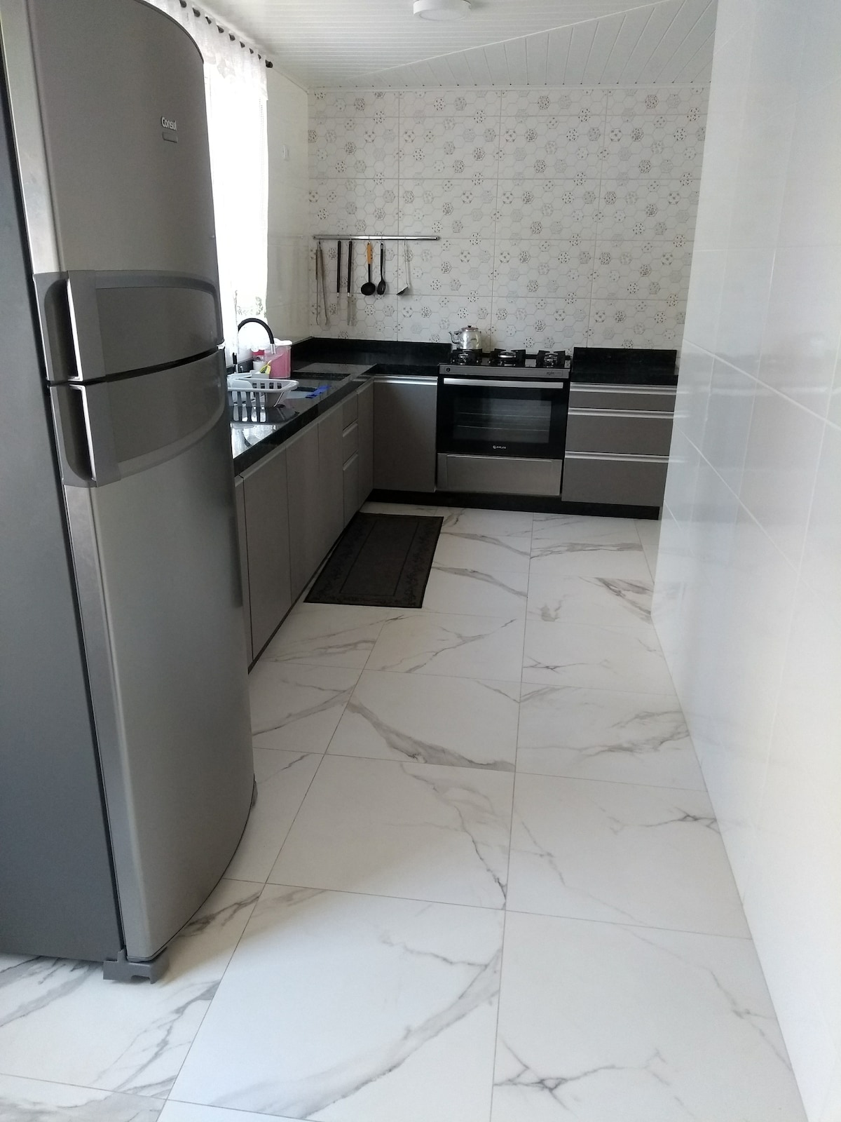 A modern kitchen is displayed with sleek cabinetry and granite countertops. Stainless steel appliances, including a refrigerator and stove, are arranged neatly. The floor features light-colored, marbled tiles, while patterned wallpaper complements the interior design. Cooking utensils are hung on a rack above the countertop.