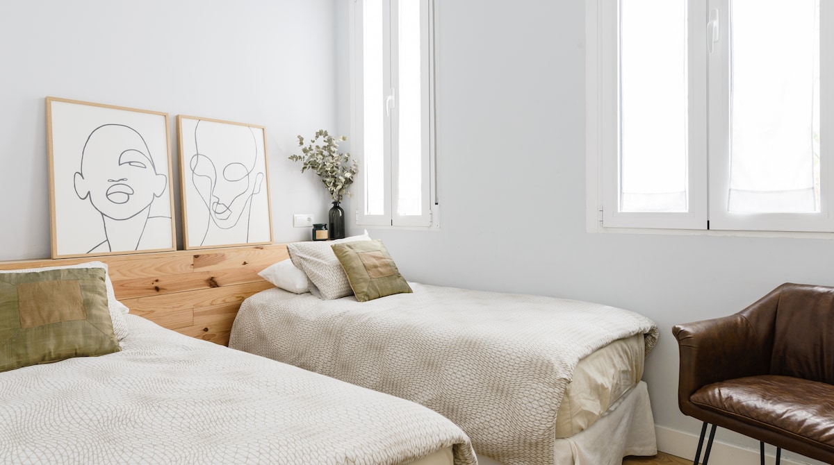 The room features two individual beds with neutral-colored bedding. A wooden headboard adds warmth, while minimalist artwork hangs on the wall above. Natural light enters through the windows, and a stylish leather chair is positioned nearby for relaxation.
