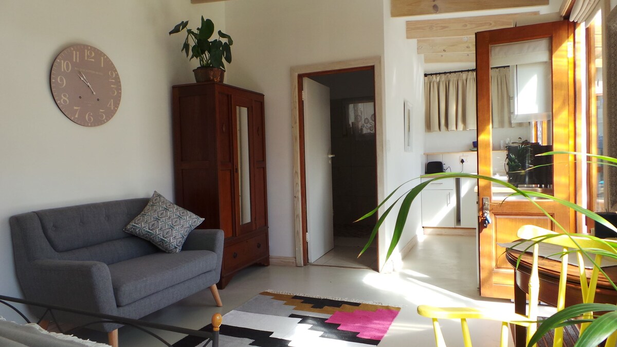 A cozy living space features a soft gray sofa and a decorative throw pillow. A wooden armoire stands against the wall, while a colorful area rug adds warmth to the floor. Natural light fills the room through large windows, enhancing the inviting ambiance.