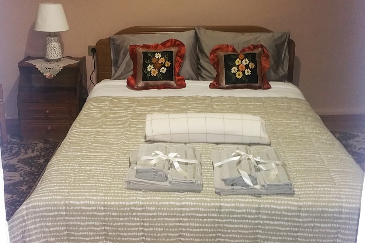 A spacious bed is adorned with decorative pillows featuring floral designs. A light-colored blanket is neatly placed at the foot of the bed. Two sets of rolled towels are arranged on the patterned carpet, adding to the room's inviting appearance. A bedside table includes a lamp.