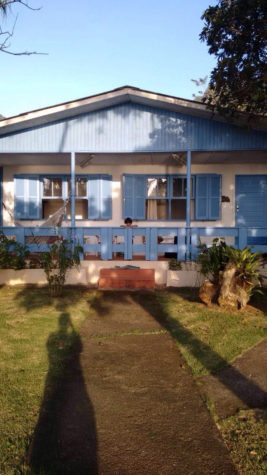 A welcoming house exterior is showcased with blue window shutters and a porch. Lush greenery lines the pathway leading to the entrance, creating a comfortable atmosphere. The building is illuminated by natural light, highlighting its charming design.
