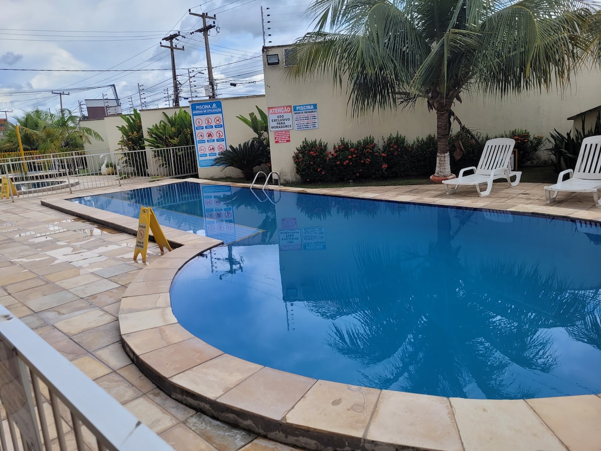 A clean swimming pool is surrounded by a tiled patio and lush greenery, providing a relaxing atmosphere. Several lounge chairs are positioned near the water's edge. Caution signs indicate safety instructions, and the pool's surface reflects the clear sky above.