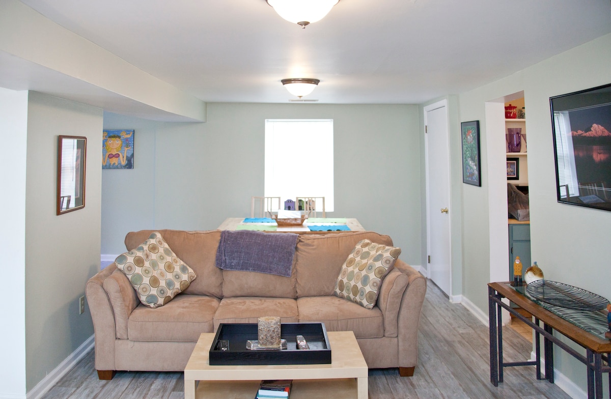 A cozy family room features a comfortable beige sofa adorned with decorative cushions. A wooden coffee table is positioned in front, and a dining area with a light wooden table and chairs is visible in the background. Soft natural light enters through a window.