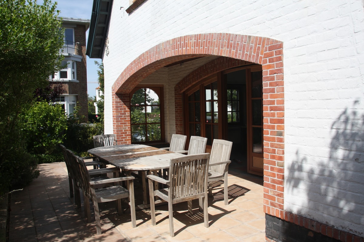 An outdoor terrace features a large wooden dining table surrounded by six matching chairs. The area is shaded by an overhanging roof, with a brick wall offering a contrast to the white facade. Lush greenery surrounds the space, enhancing the inviting setting.