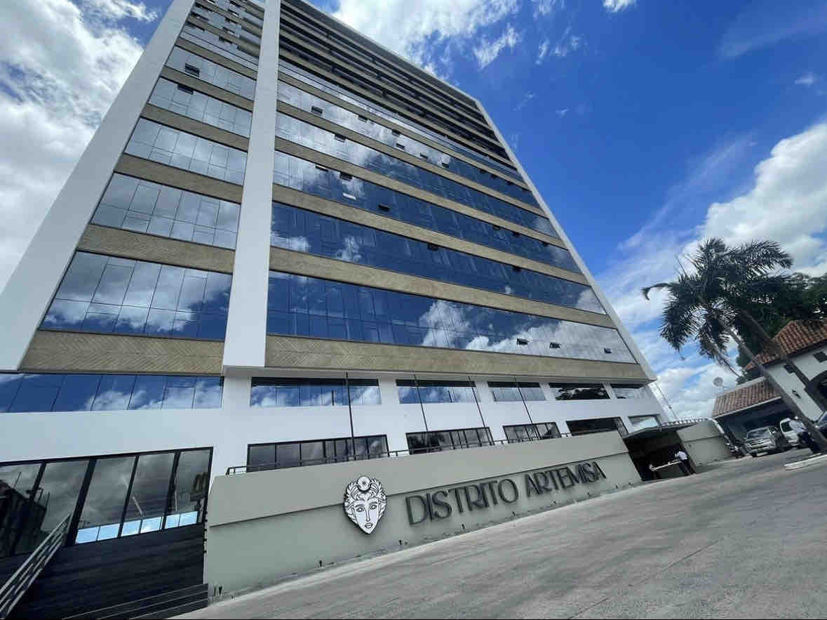 The modern tower of Distrito Artemisa is presented against a backdrop of blue sky, showcasing large glass windows reflecting the surroundings. The entrance features a sleek design with a prominent sign, inviting guests to explore the contemporary architecture of this central location.