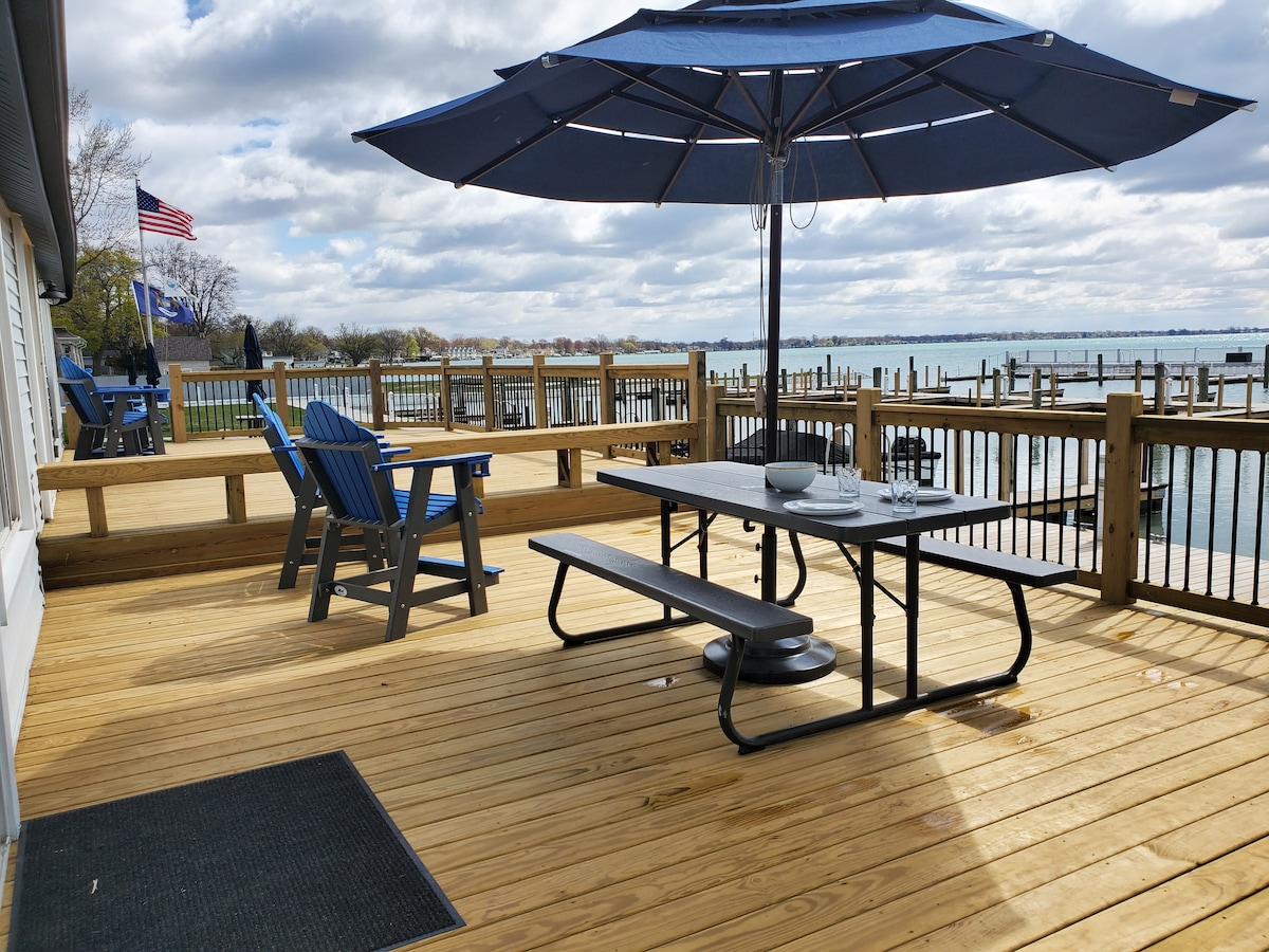 A spacious outdoor deck features a dark blue umbrella providing shade over a black picnic table. Blue Adirondack chairs surround the area, with a view of the water and wooden docks in the background. A welcoming atmosphere is created by the natural wood flooring.