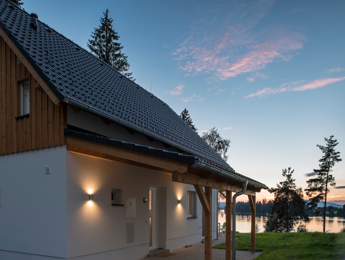 A modern house exterior is viewed at dusk, featuring a sloped roof with dark tiles. Soft lighting highlights the entrance, while scenic views of a lake and trees are visible in the background, creating a serene atmosphere.