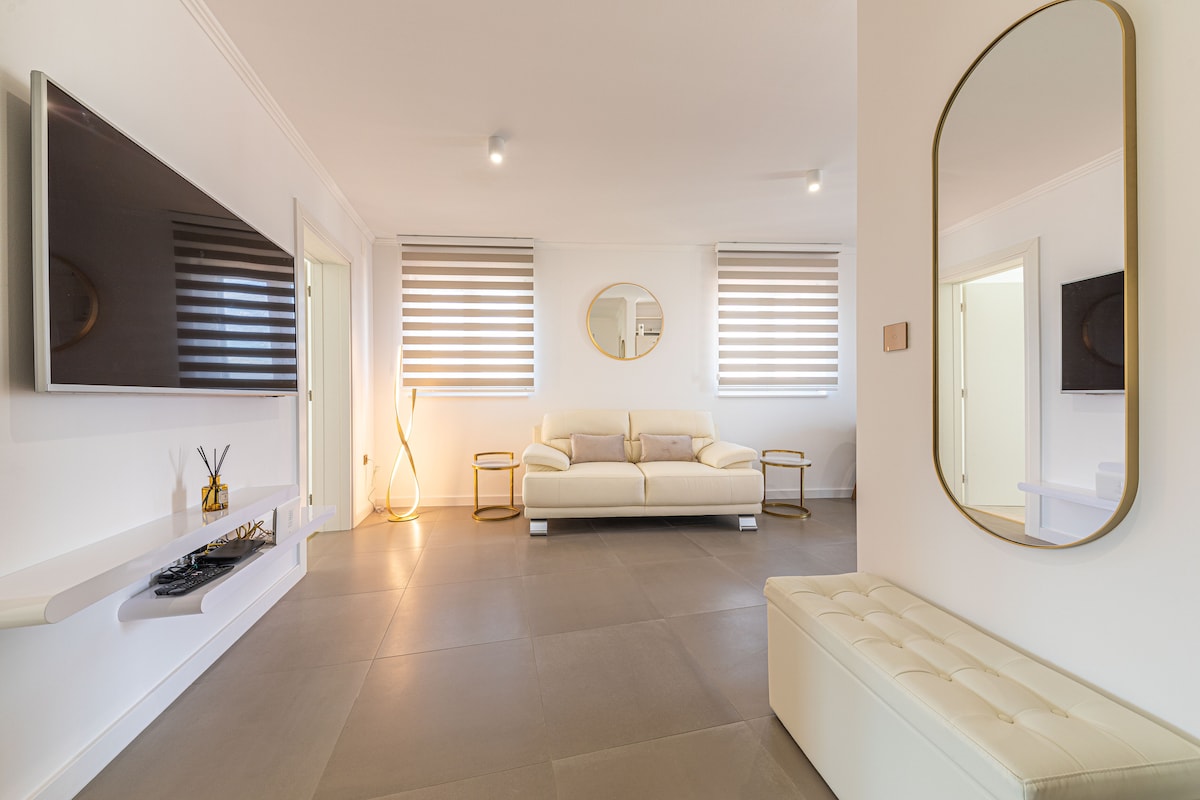 A spacious living area features a light-toned leather sofa facing dual flat-screen televisions mounted on the wall. Decorative mirrors enhance the room's brightness. Soft, ambient lighting highlights modern furniture, while window coverings allow natural light to filter softly into the space.