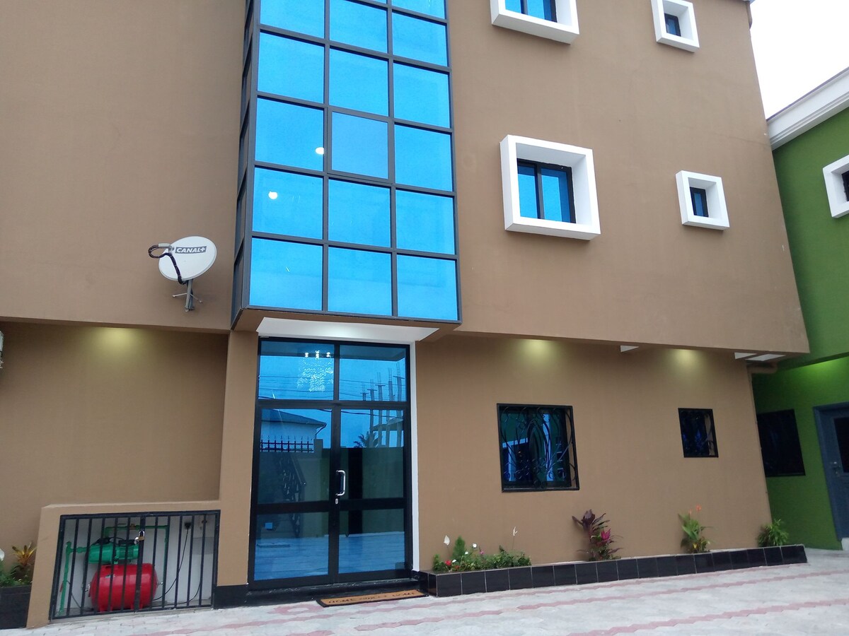 The exterior of the residence features a modern design with large blue glass windows and a brown facade. A welcoming entrance is highlighted by a glass door, with multiple smaller windows providing natural light. Decorative plants are positioned along the front.