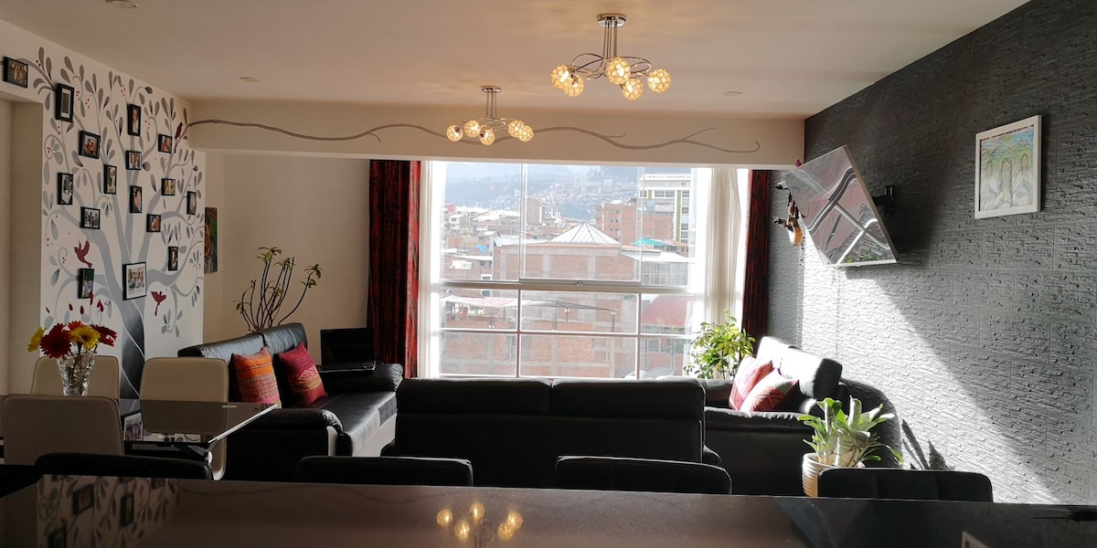 A modern living area is equipped with a sectional sofa and a small table, complemented by vibrant decor and artwork on the walls. Large windows provide natural light and showcase a view of the city and mountains. Contemporary light fixtures hang from the ceiling.