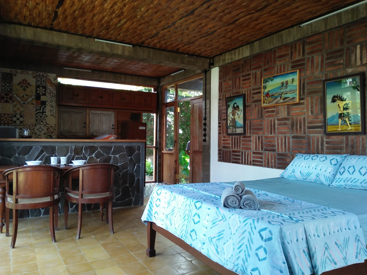 A spacious cottage interior features a large bed with a patterned blanket and neatly arranged towels. A wooden dining table with chairs is placed near the entrance, while artwork adorns the exposed brick wall. A kitchenette is visible in the background.