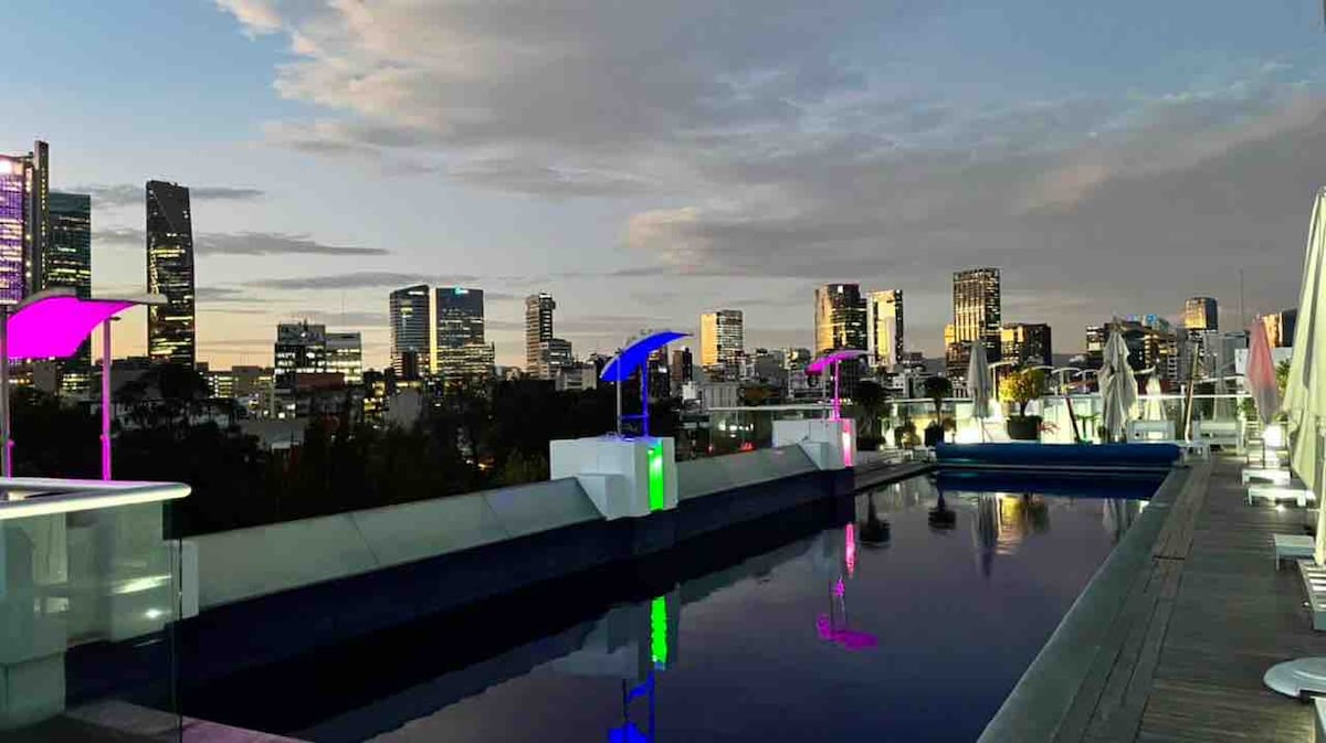A rooftop pool area is showcased with an infinity edge, overlooking a city skyline at twilight. Colorful illuminated umbrellas provide seating, complemented by modern lounge chairs. The tranquil pool reflects the evening sky, offering a serene urban retreat.