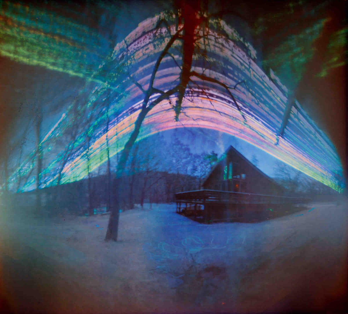 A wooden chalet sits nestled in a serene landscape, surrounded by snow-covered ground and bare trees. Clear skies reflect light above, creating a colorful effect. The structure features a sloped roof with visible windows, providing an inviting presence amidst nature.