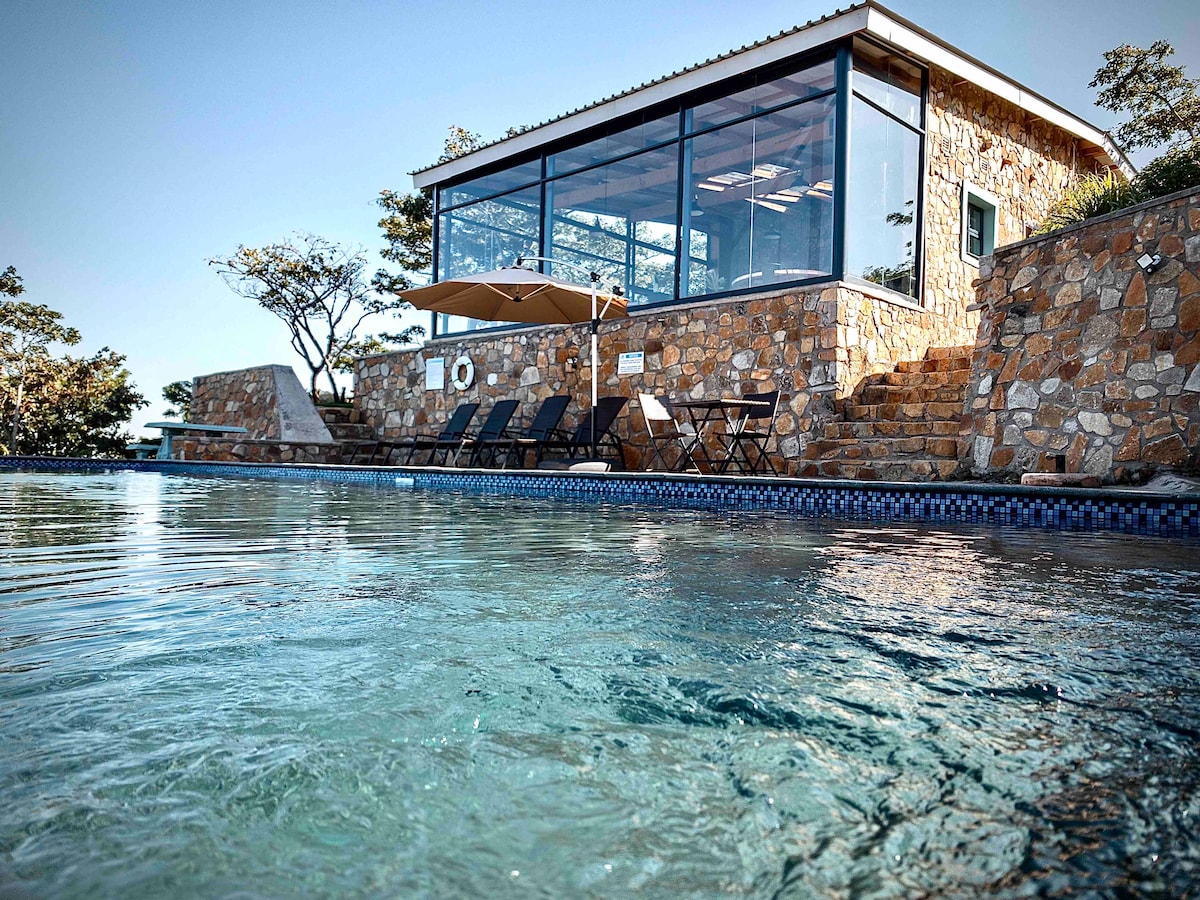 A stone structure reflects rustic charm, featuring large glass windows that allow natural light to fill the interior. A shaded area with lounge chairs is positioned beside a sparkling pool, creating a serene atmosphere perfect for relaxation.