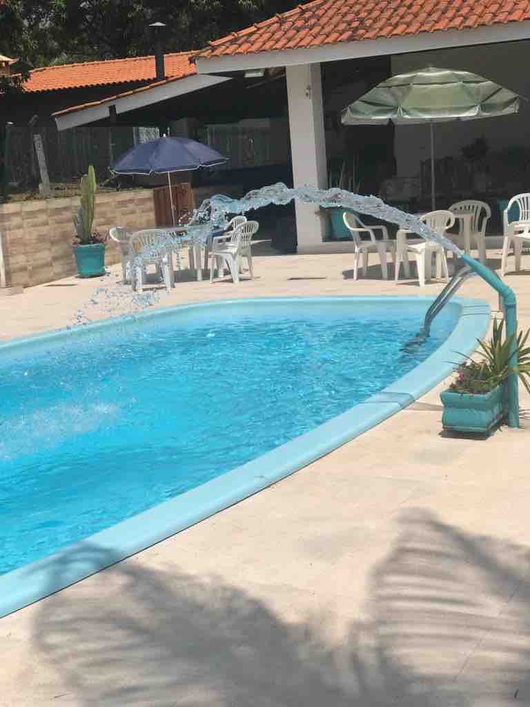 A refreshing swimming pool is featured, with water cascading from a decorative fountain. Surrounding the pool are multiple white chairs and tables, providing ample seating. A shaded area with umbrellas is present, offering a comfortable retreat. Green plants add a touch of nature to the space.