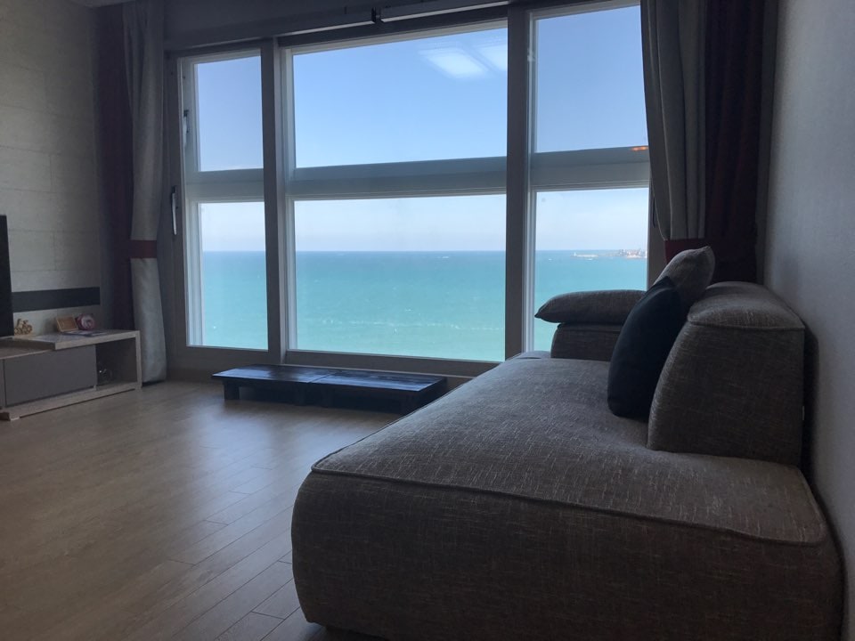 A spacious seating area features a large window showcasing a panoramic view of the open sea. Natural light floods in, highlighting aspects of the room, while a cozy grey sofa offers a comfortable spot to take in the scenery.