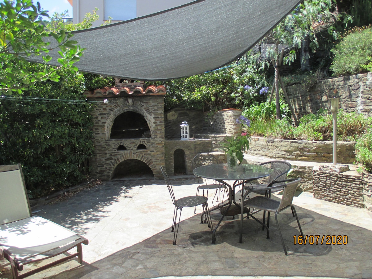 An outdoor terrace is depicted, featuring a stone barbecue and a round table with metal chairs. Shade is provided by a canopy overhead. Surrounding greenery adds privacy, while a stone wall forms part of the background, enhancing the outdoor space's natural feel.