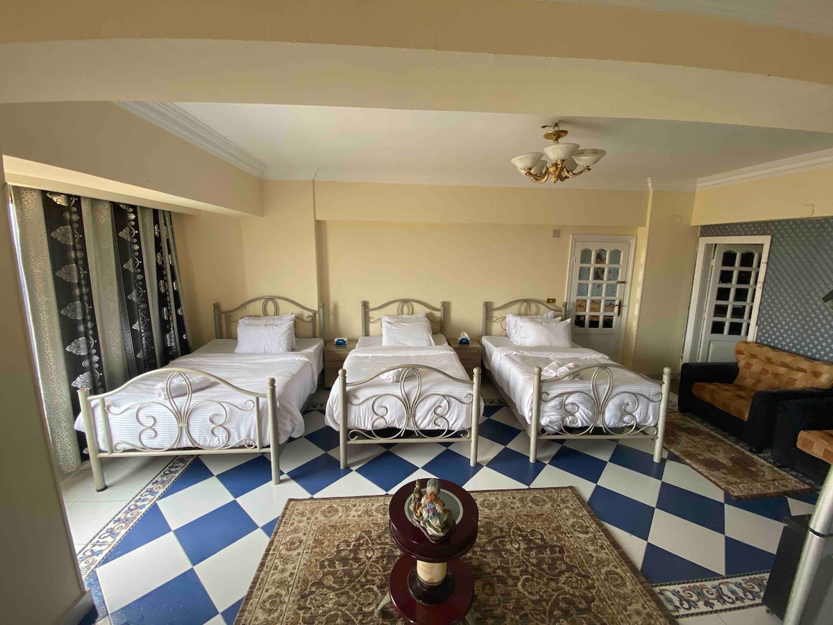 A spacious room is presented with three beds arranged neatly, each made up with white linens. A round table is placed centrally, adorned with decorative items. Natural light filters through large windows, and the floor features a patterned tile design, complementing the overall decor.
