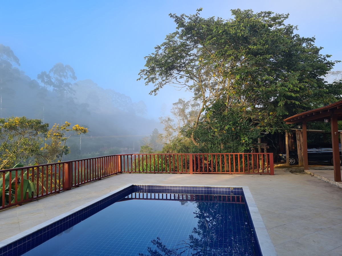 A serene pool area is visible, surrounded by a wooden railing and lush greenery. Fog envelops the distant trees, creating a tranquil atmosphere. The pool reflects the gentle morning light, harmonizing with the natural landscape.