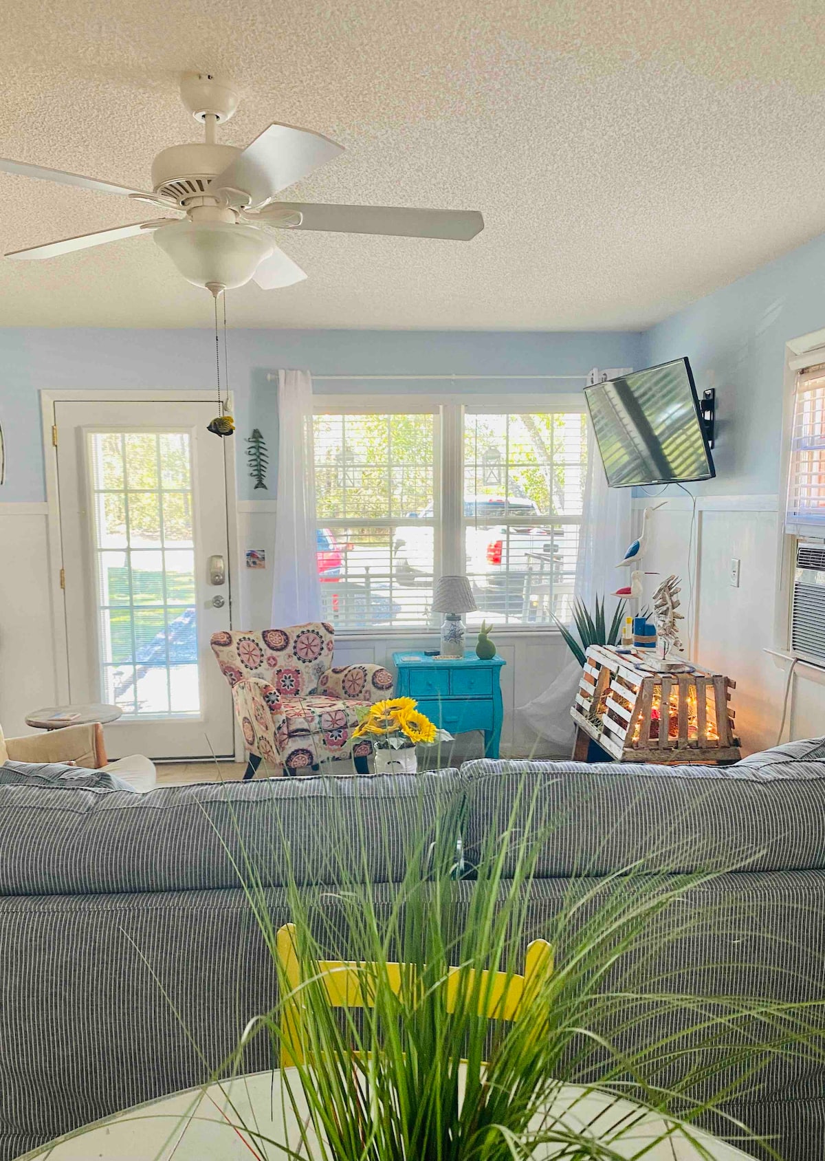 The living area features a comfortable seating arrangement with colorful chairs and a table adorned with a plant. Large windows allow natural light to fill the space, complemented by light blue walls and a ceiling fan. A television and decorative elements enhance the welcoming ambiance.