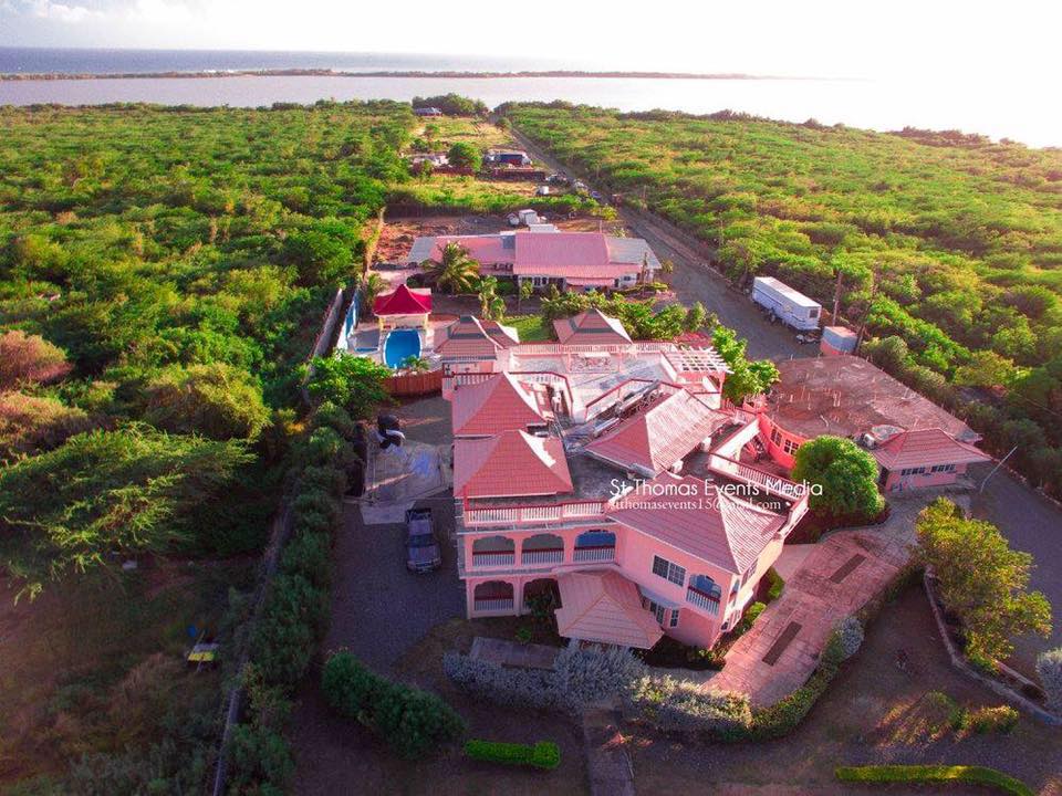 An aerial view of Casa Lagoona Hotel reveals a spacious property with vibrant pink buildings surrounded by lush greenery. The hotel features a pool, garden, and multiple outdoor spaces, with the tranquil Yallahs Lagoon visible in the background under a soft sunset glow.
