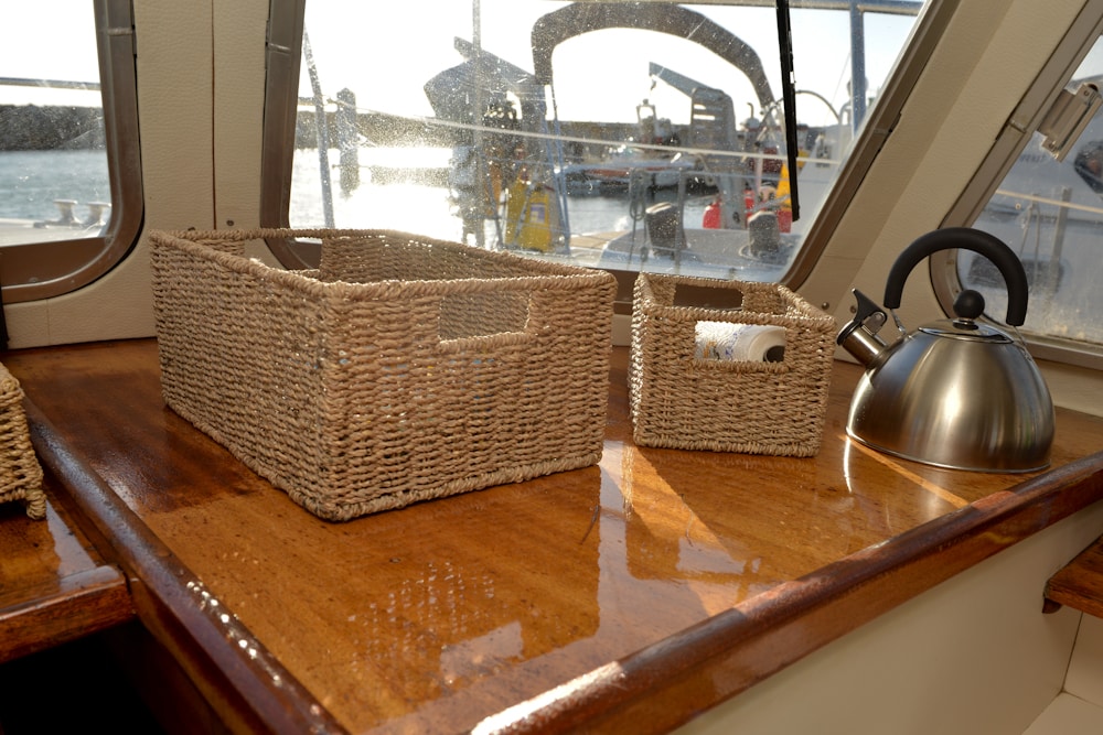 A polished wooden table features woven storage baskets, one larger and one smaller. A stainless steel kettle is positioned nearby. Sunlight streams through the windows, creating a warm and inviting atmosphere while a view of the marina is visible outside.